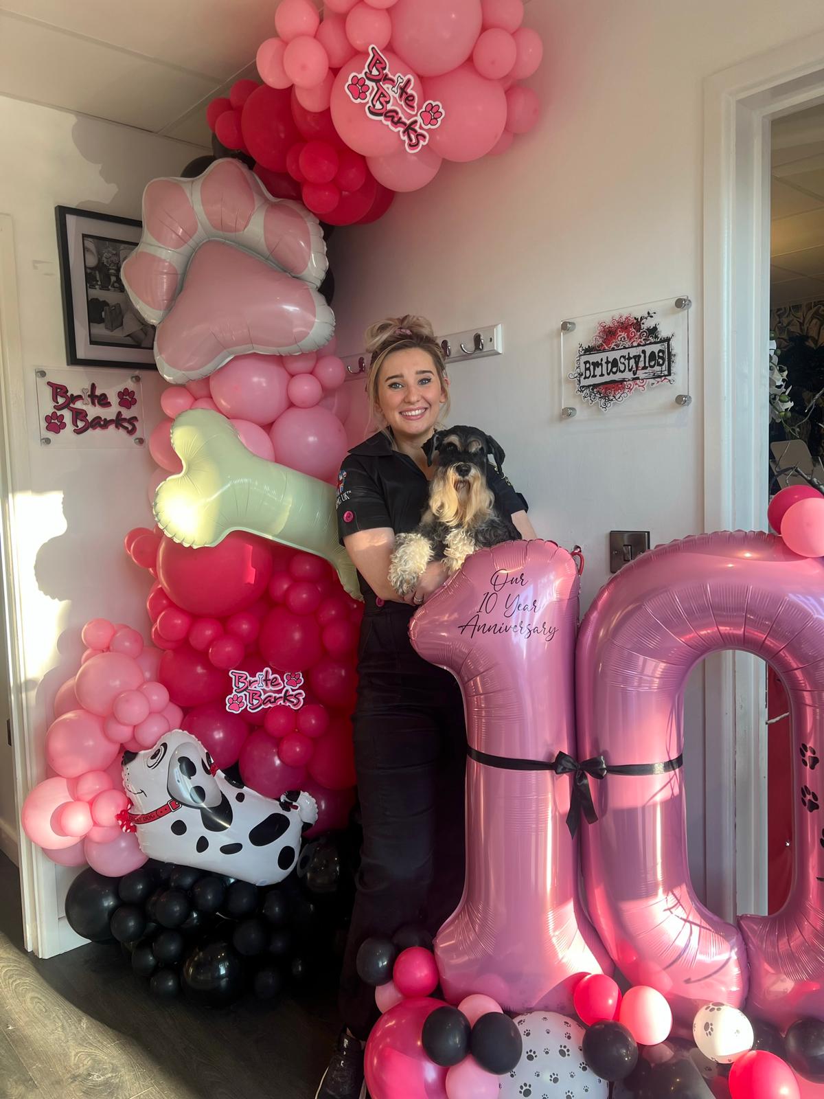 A groomer holding a puppy beside a pink balloon arch in the salon